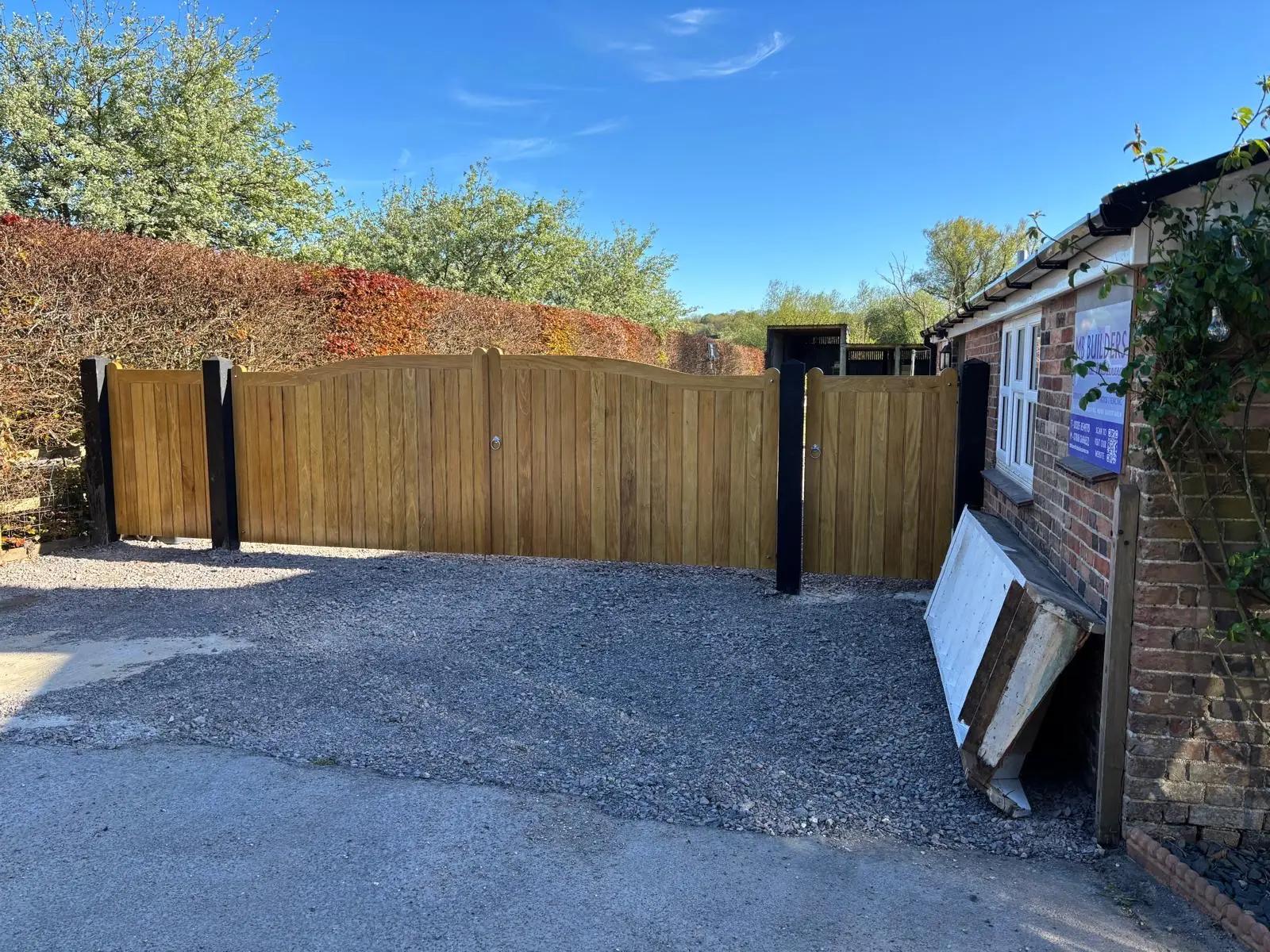Curved-top softwood timber driveway gates on black posts beside a brick outbuilding