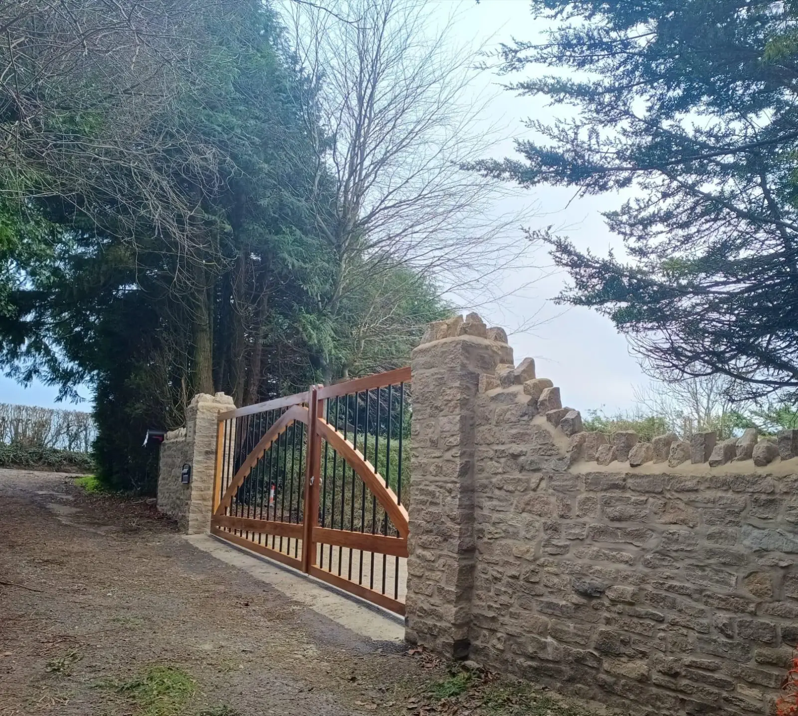 Timber driveway gate with arched bracing set into a curved dry-stone wall on a wooded lane